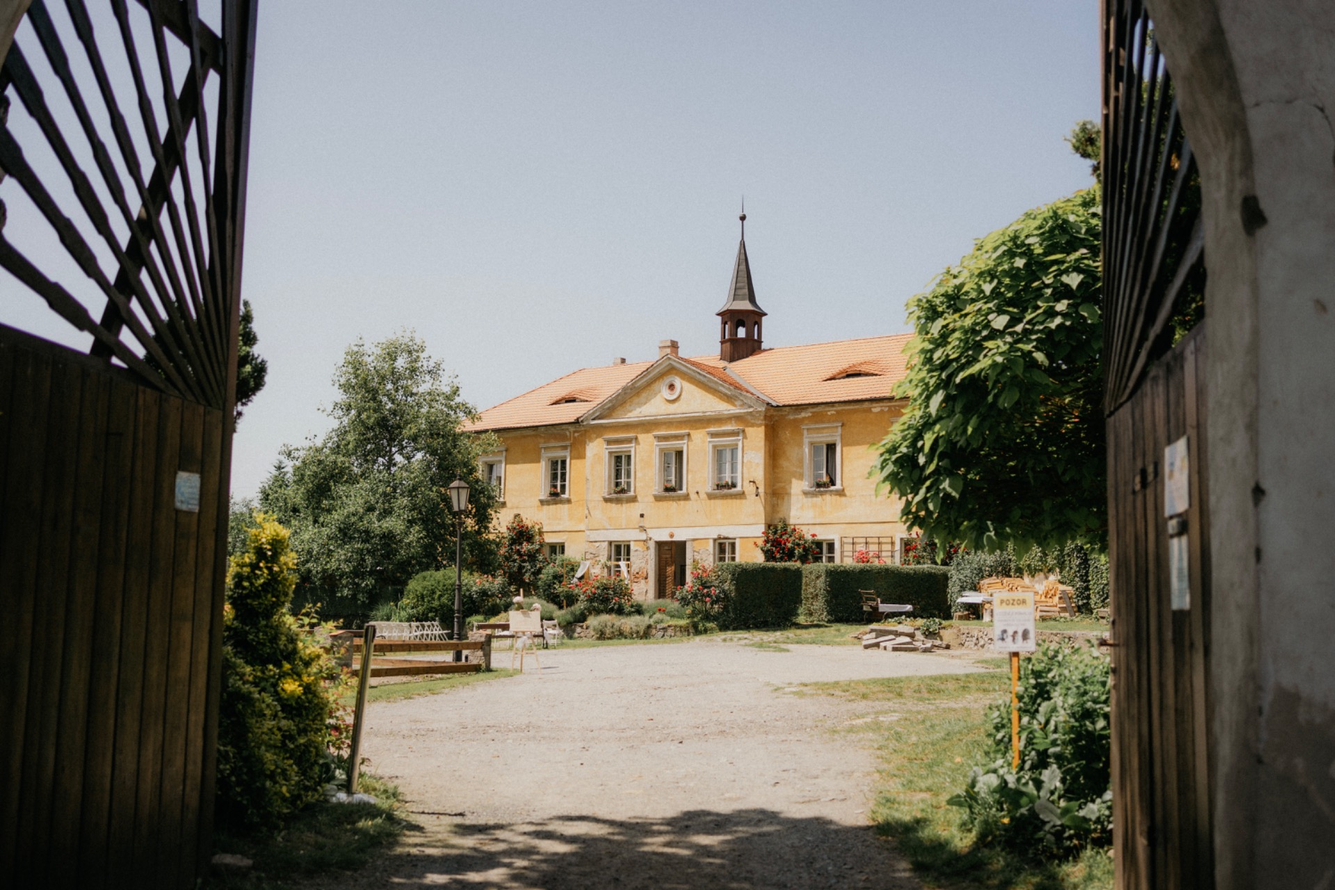 Novomanželé stojí u historické fasády s květinovou výzdobou, zelené věnce a červené muškáty v pozadí.